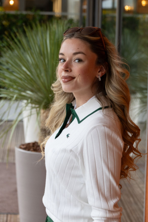 Side profile of Emma Broyon displaying voluminous blonde waves and classic Lacoste white ribbed polo with green trim at Roland Garros 2025