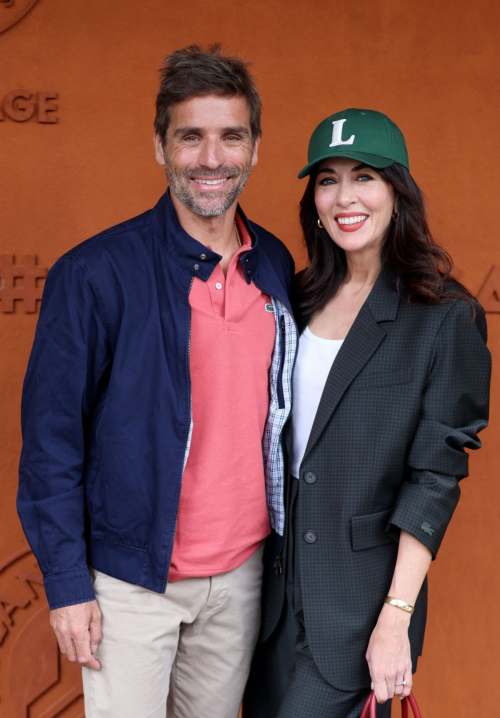 Nolwenn Leroy posing with companion at Roland Garros 2025 wearing elegant dark blazer outfit and green Lacoste cap against terracotta backdrop