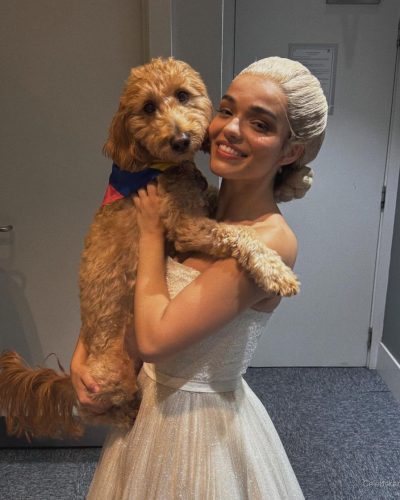 Rachel Zegler in white dress with playful dog beside her.