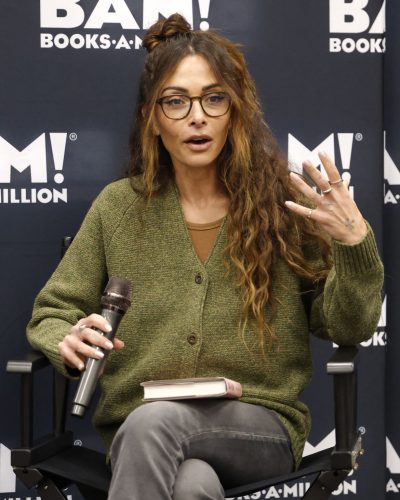 Sarah Shahi styled in a warm green cardigan with gray trousers.