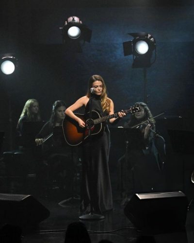 Maggie Rogers in minimal black dress with dramatic lighting backdrop.