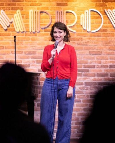 Milana Vayntrub in bright red cardigan and high-waisted blue jeans.