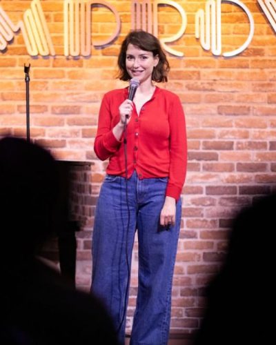 Milana Vayntrub wearing a red cardigan and comfortable blue jeans.